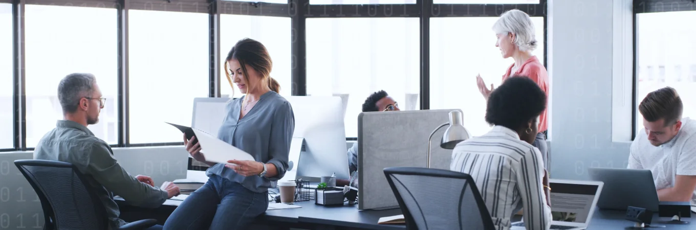 Sei persone lavorano in un ufficio luminoso, chiacchierando, leggendo o scrivendo al computer.