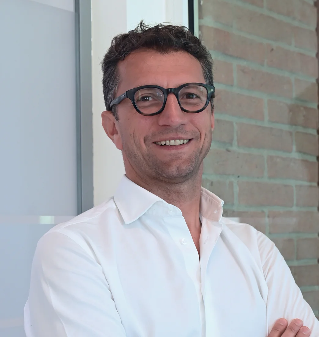 Un uomo sorridente con gli occhiali e una camicia bianca è in piedi accanto a un muro di mattoni.