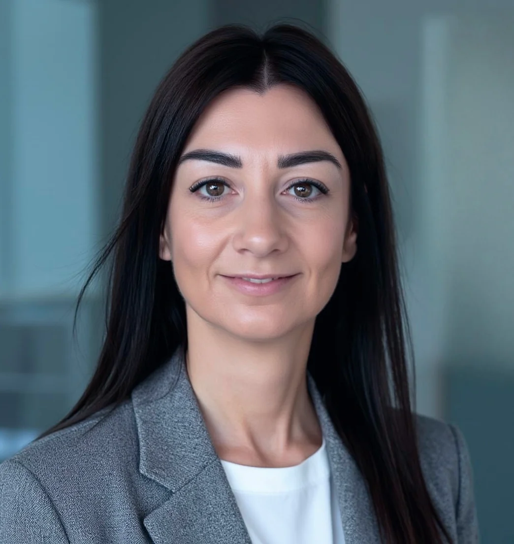 Una donna con un blazer grigio è in piedi in un ufficio e guarda la telecamera con espressione neutra.